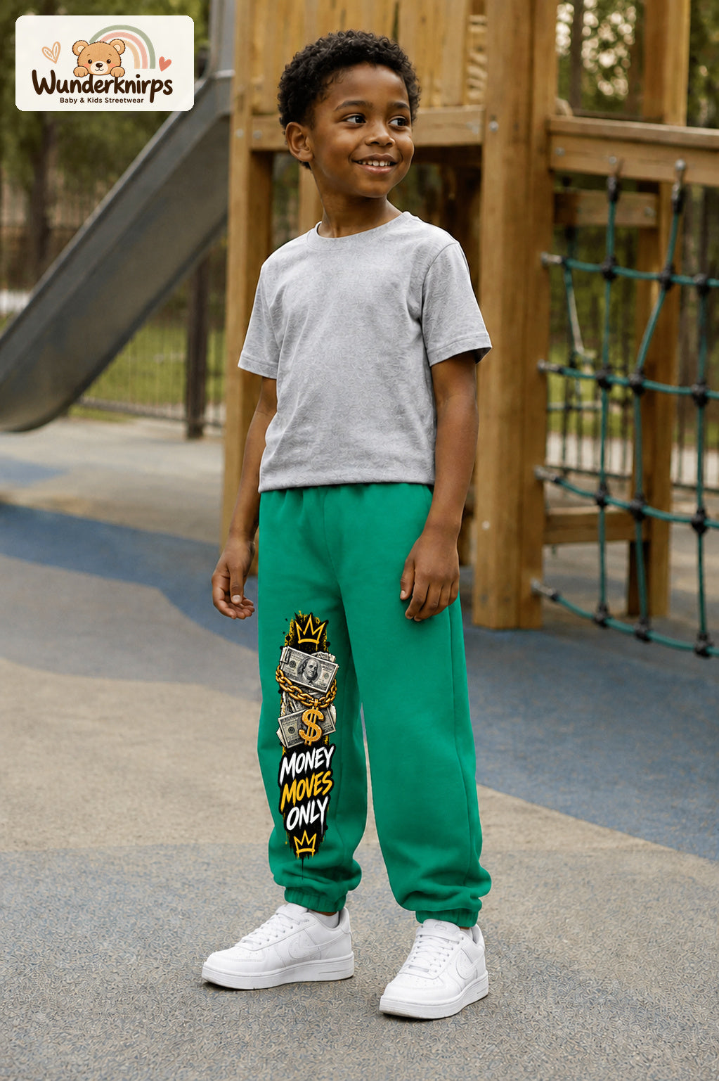 Junge trägt grüne Jogginghose mit Money Moves Gold-Print, leicht seitlich auf dem Spielplatz.