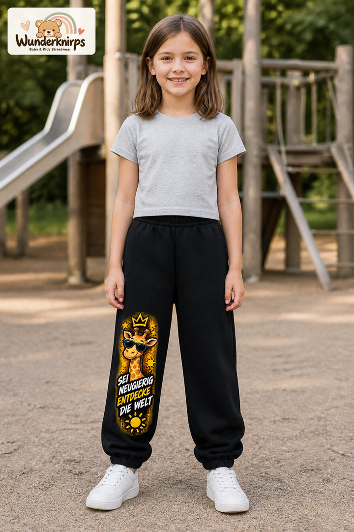 Mädchen trägt schwarze Jogginghose mit Neugierig Giraffe-Print, von vorn auf dem Spielplatz.