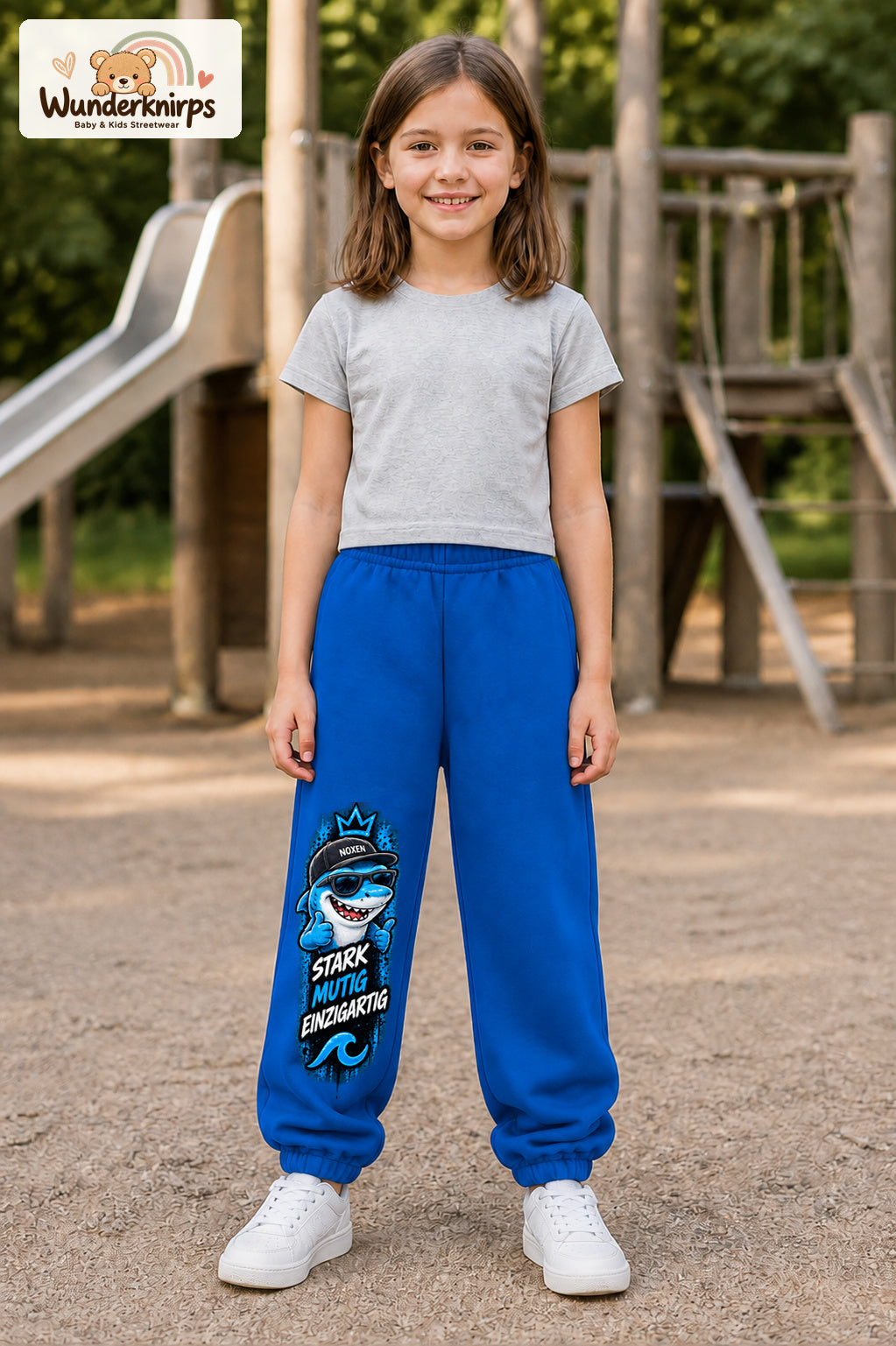 Mädchen trägt blaue Jogginghose mit Stark einzigartig Cap-Hai-Print, von vorn auf dem Spielplatz.
