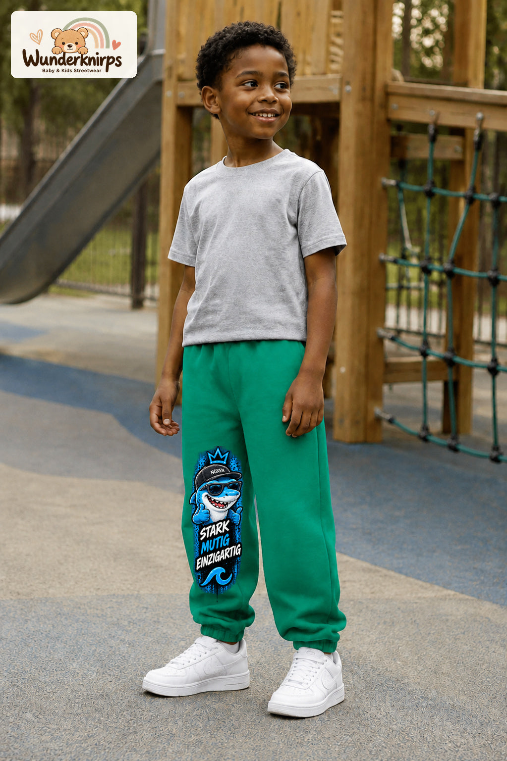 Junge trägt grüne Jogginghose mit Stark einzigartig Cap-Hai-Print, leicht seitlich auf dem Spielplatz.