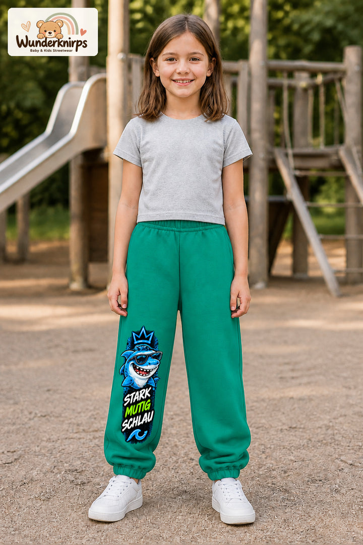 Mädchen trägt grüne Jogginghose mit Stark mutig schlau Hai-Print, von vorn auf dem Spielplatz.