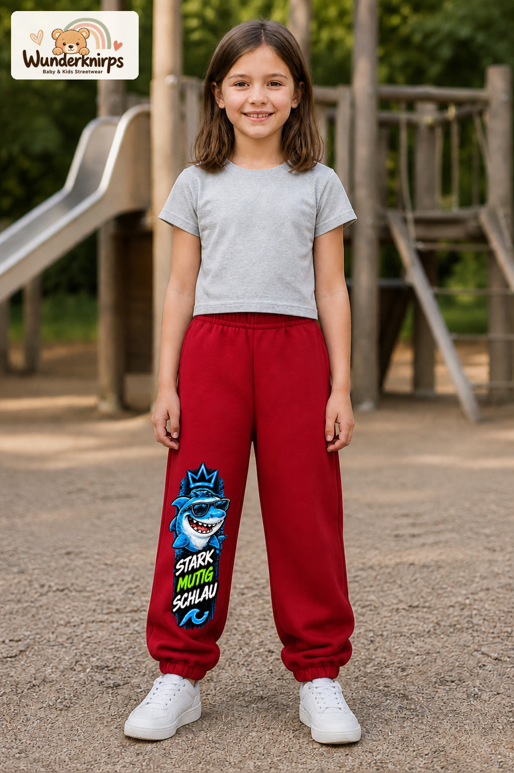 Mädchen trägt rote Jogginghose mit Stark mutig schlau Hai-Print, von vorn auf dem Spielplatz.