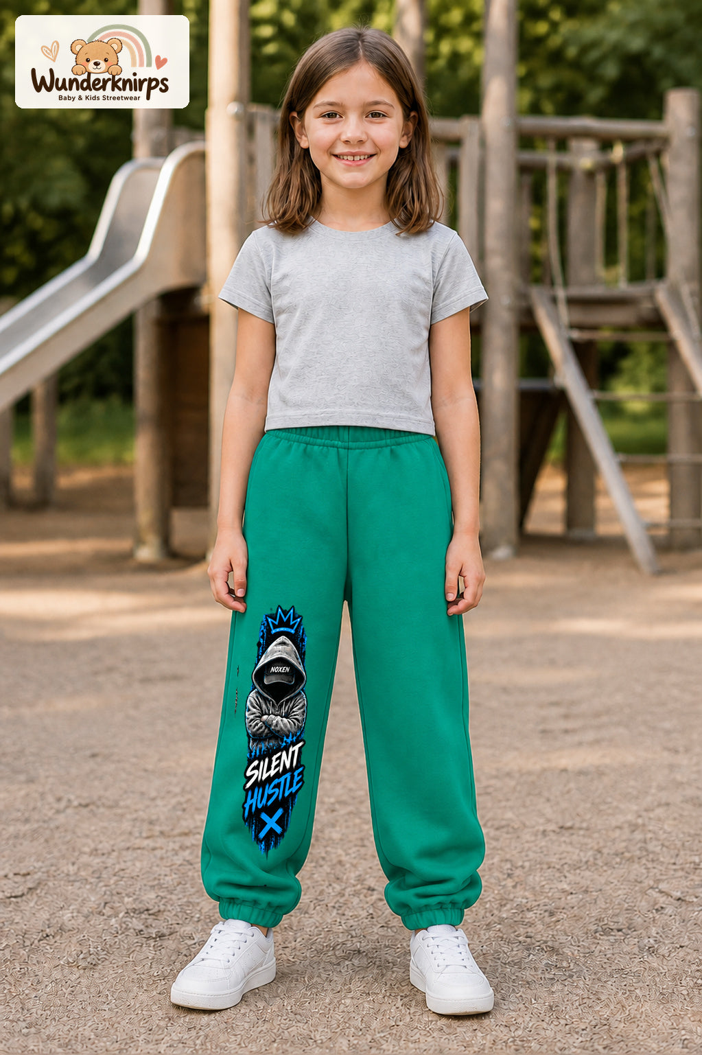 Mädchen trägt grüne Jogginghose mit Silent Hustle Hoodie-Print, von vorn auf dem Spielplatz.