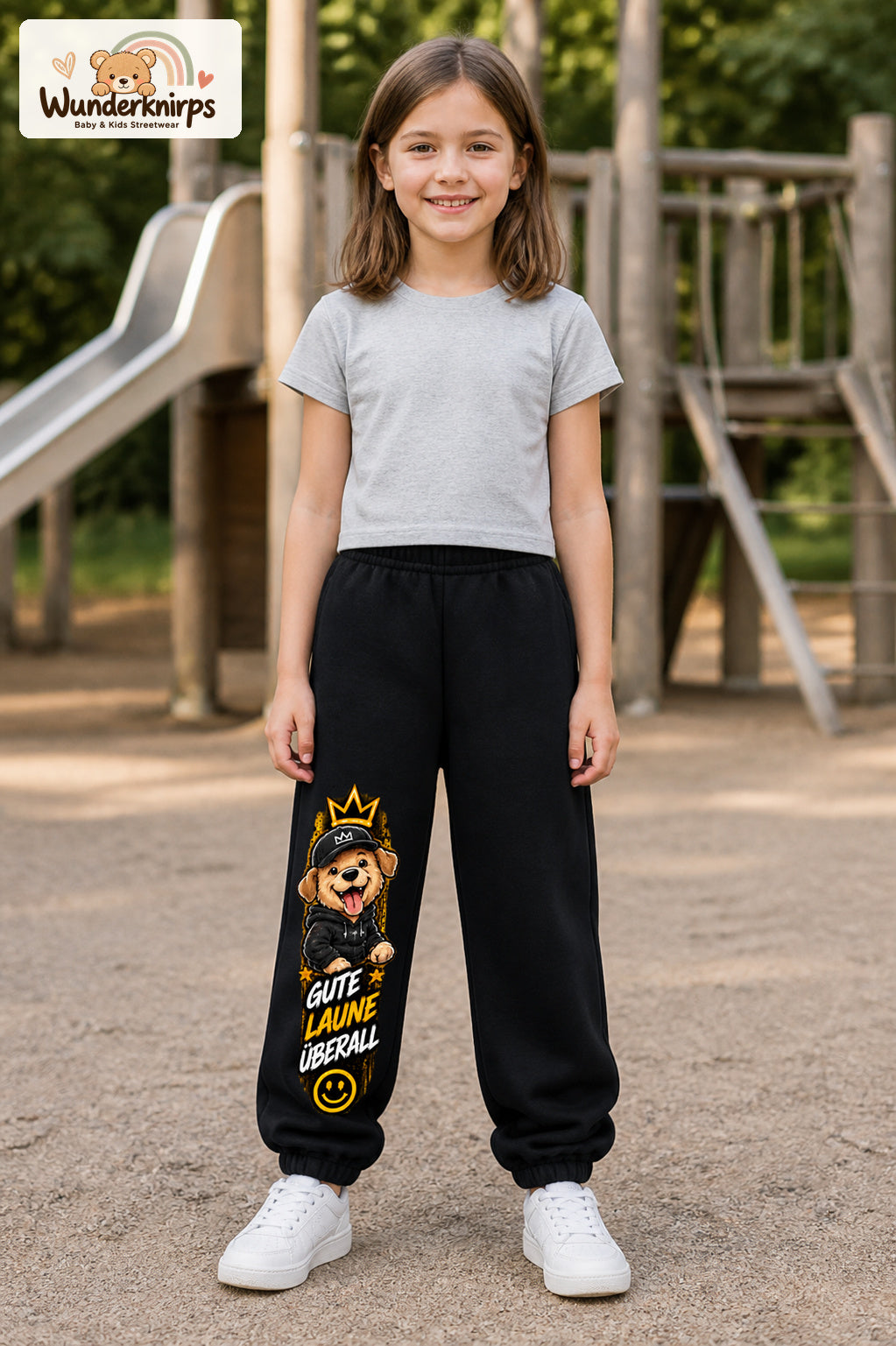 Mädchen trägt schwarze Jogginghose mit Gute Laune Cap-Hund-Print, von vorn auf dem Spielplatz.