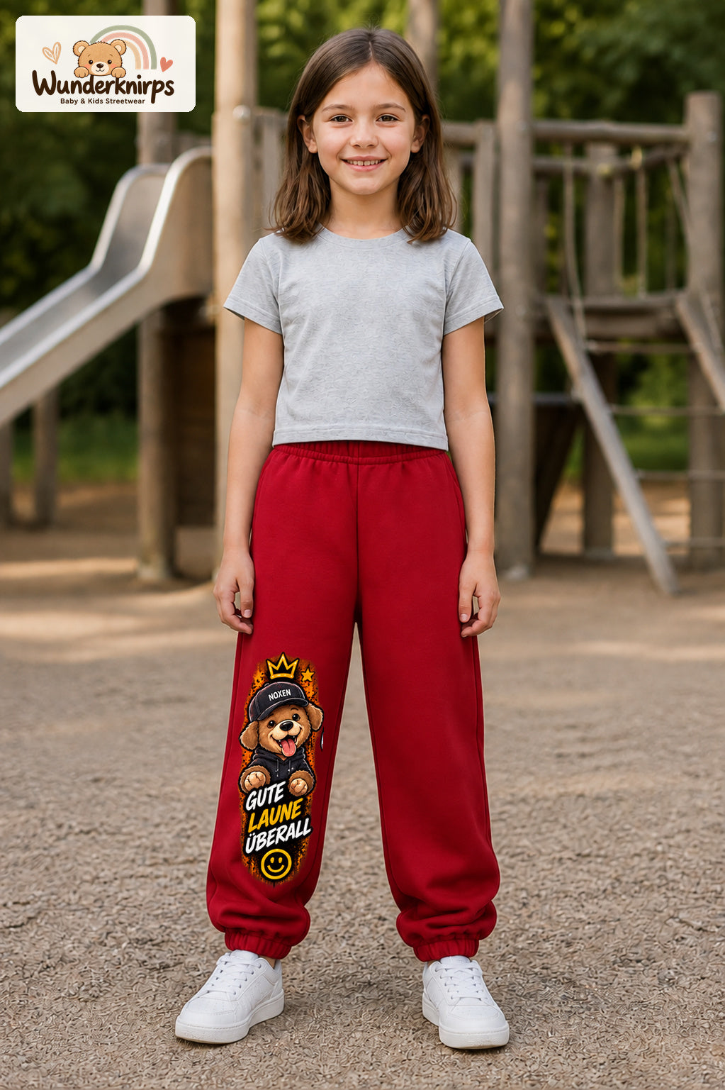 Mädchen trägt rote Jogginghose mit Gute Laune Hoodie-Hund-Print, von vorn auf dem Spielplatz.