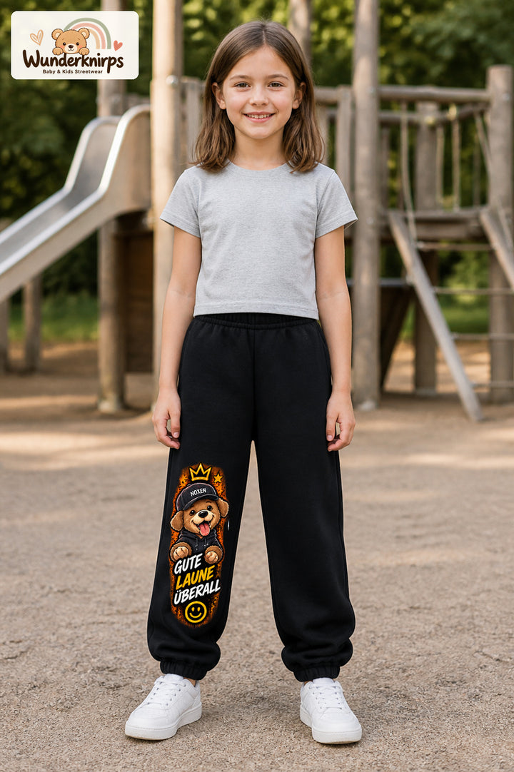 Mädchen trägt schwarze Jogginghose mit Gute Laune Hoodie-Hund-Print, von vorn auf dem Spielplatz.