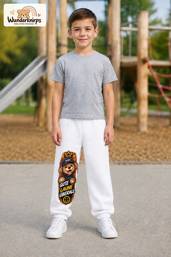 Junge trägt weiße Jogginghose mit Gute Laune Hoodie-Hund-Print, von vorn auf dem Spielplatz.