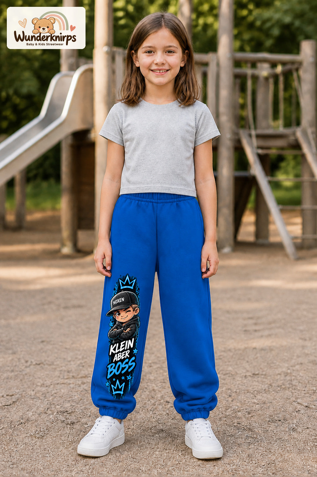 Mädchen trägt blaue Jogginghose mit Klein aber Boss Cap-Print, von vorn auf dem Spielplatz.