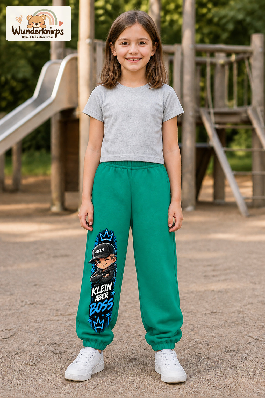 Mädchen trägt grüne Jogginghose mit Klein aber Boss Cap-Print, von vorn auf dem Spielplatz.
