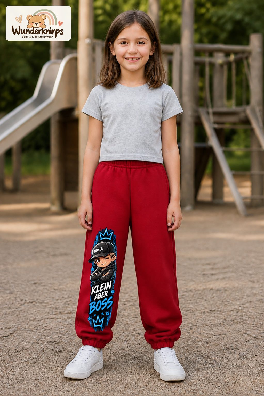 Mädchen trägt rote Jogginghose mit Klein aber Boss Cap-Print, von vorn auf dem Spielplatz.