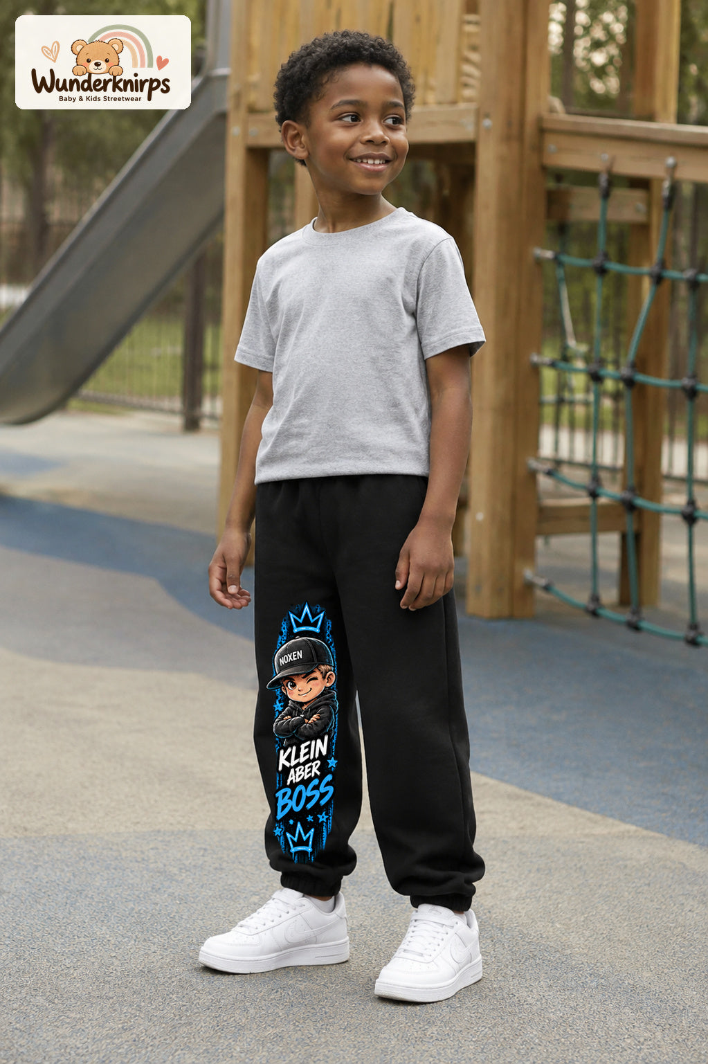 Junge trägt schwarze Jogginghose mit Klein aber Boss Cap-Print, leicht seitlich auf dem Spielplatz.