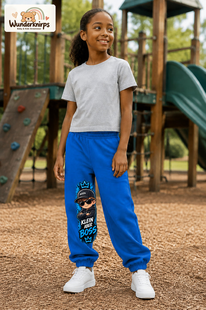 Mädchen trägt blaue Jogginghose mit Klein aber Boss Sonnenbrille-Print, leicht seitlich auf dem Spielplatz.