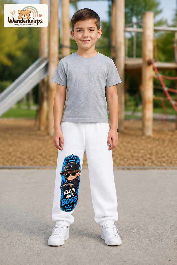 Junge trägt weiße Jogginghose mit Klein aber Boss Sonnenbrille-Print, von vorn auf dem Spielplatz.