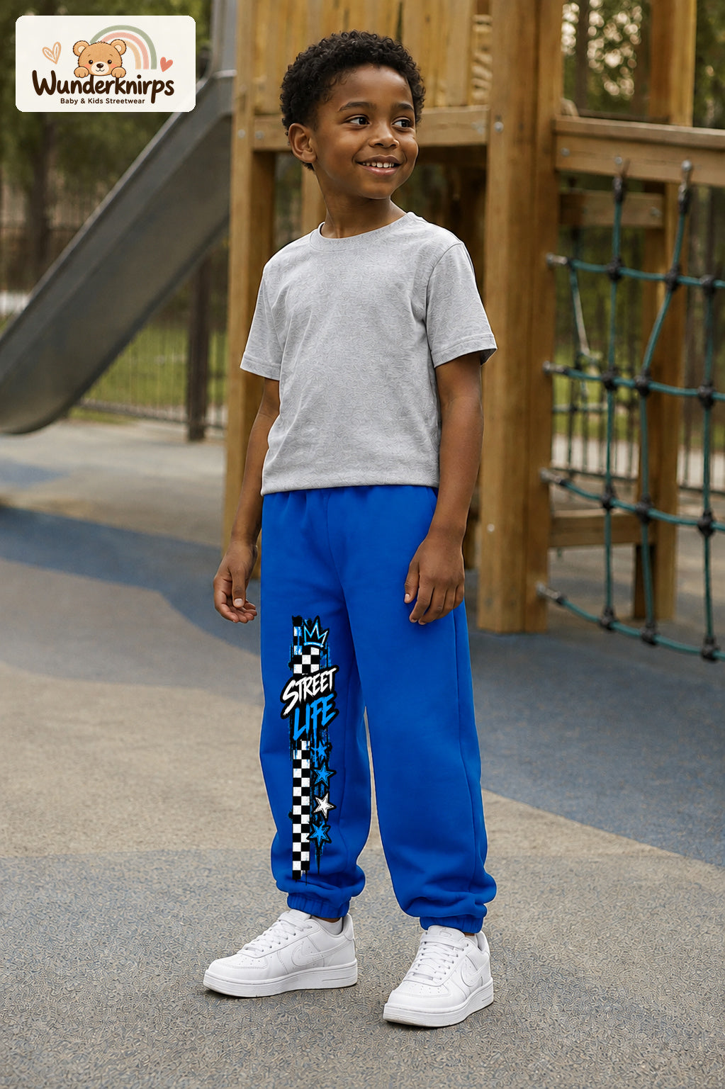 Junge trägt blaue Jogginghose mit Street Life Karo-Print, leicht seitlich auf dem Spielplatz.