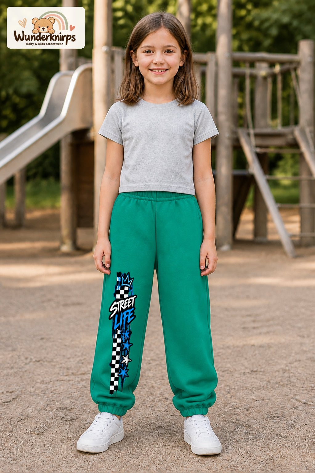 Mädchen trägt grüne Jogginghose mit Street Life Karo-Print, von vorn auf dem Spielplatz.