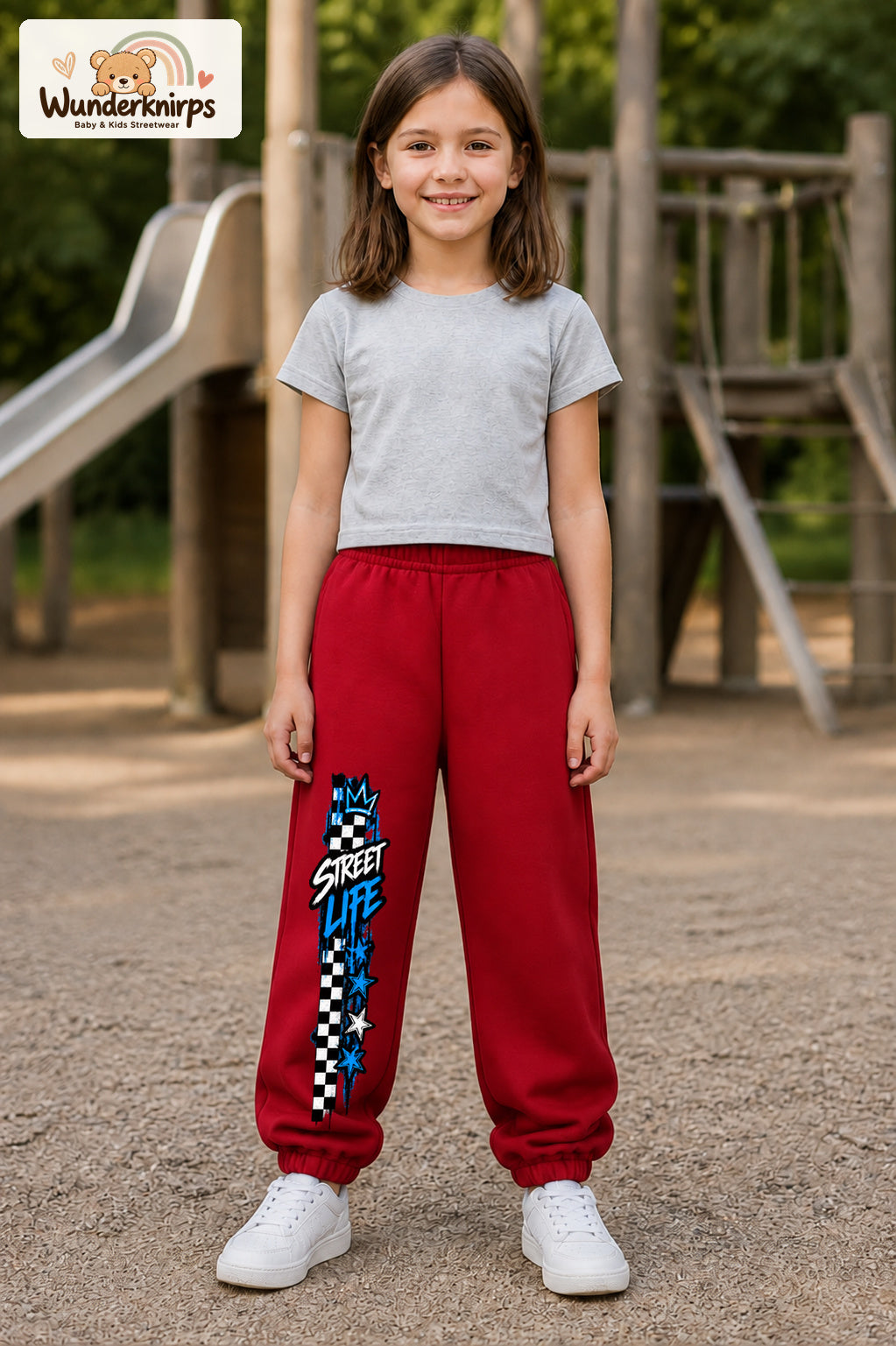 Mädchen trägt rote Jogginghose mit Street Life Karo-Print, von vorn auf dem Spielplatz.