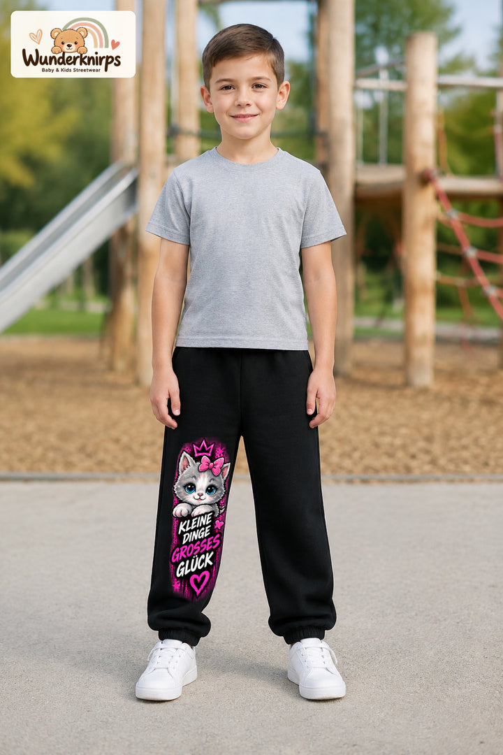 Junge trägt schwarze Jogginghose mit Großes Glück Schleifenkatze-Print, von vorn auf dem Spielplatz.