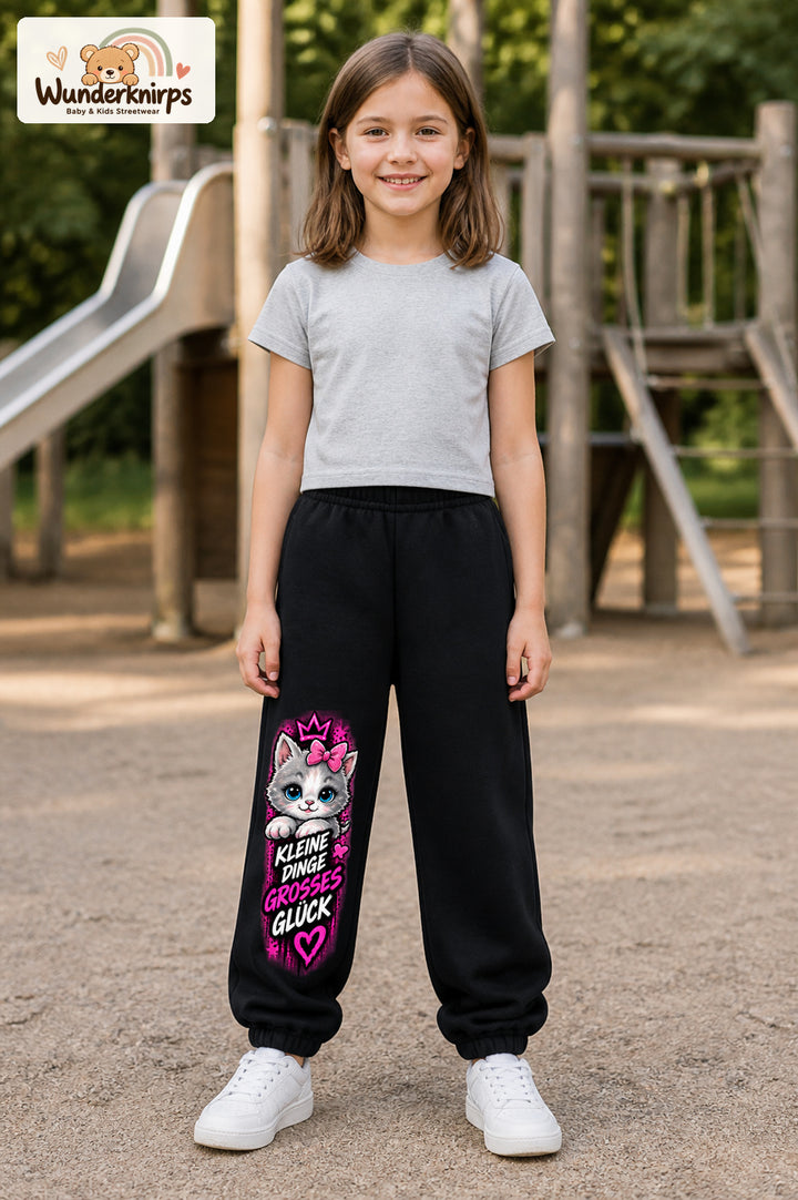 Mädchen trägt schwarze Jogginghose mit Großes Glück Schleifenkatze-Print, von vorn auf dem Spielplatz.