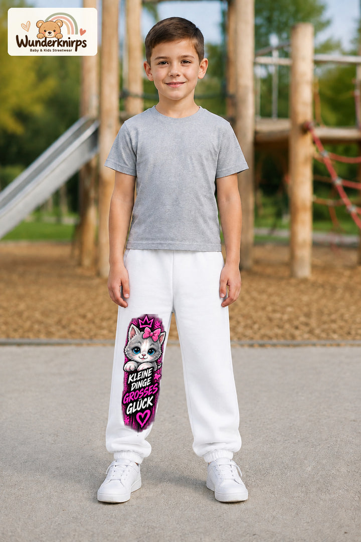 Junge trägt weiße Jogginghose mit Großes Glück Schleifenkatze-Print, von vorn auf dem Spielplatz.