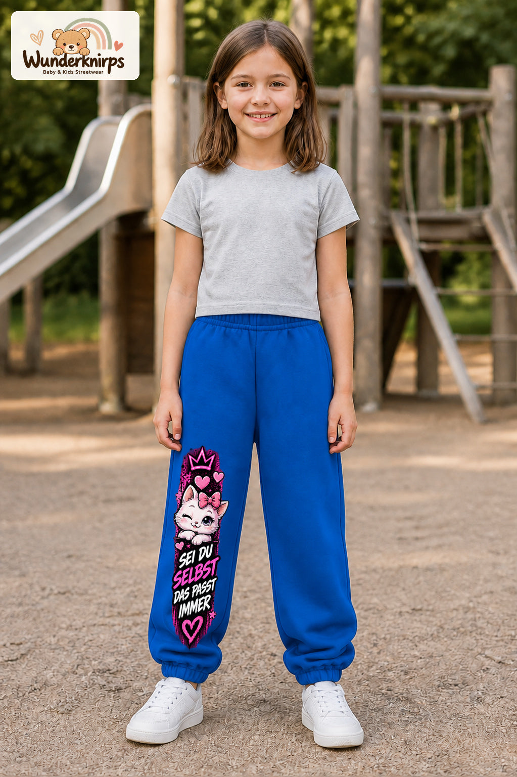 Mädchen trägt blaue Jogginghose mit Sei du selbst Kätzchen-Print, von vorn auf dem Spielplatz.
