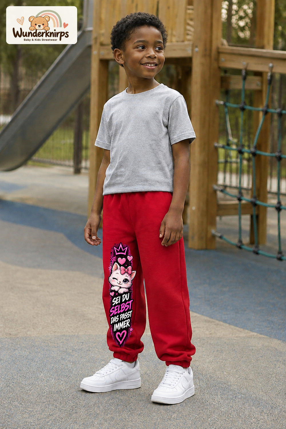 Junge trägt rote Jogginghose mit Sei du selbst Kätzchen-Print, leicht seitlich auf dem Spielplatz.