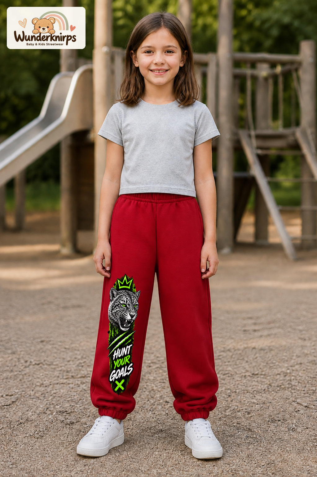 Mädchen trägt rote Jogginghose mit Hunt Your Goals Leopard-Print, von vorn auf dem Spielplatz.