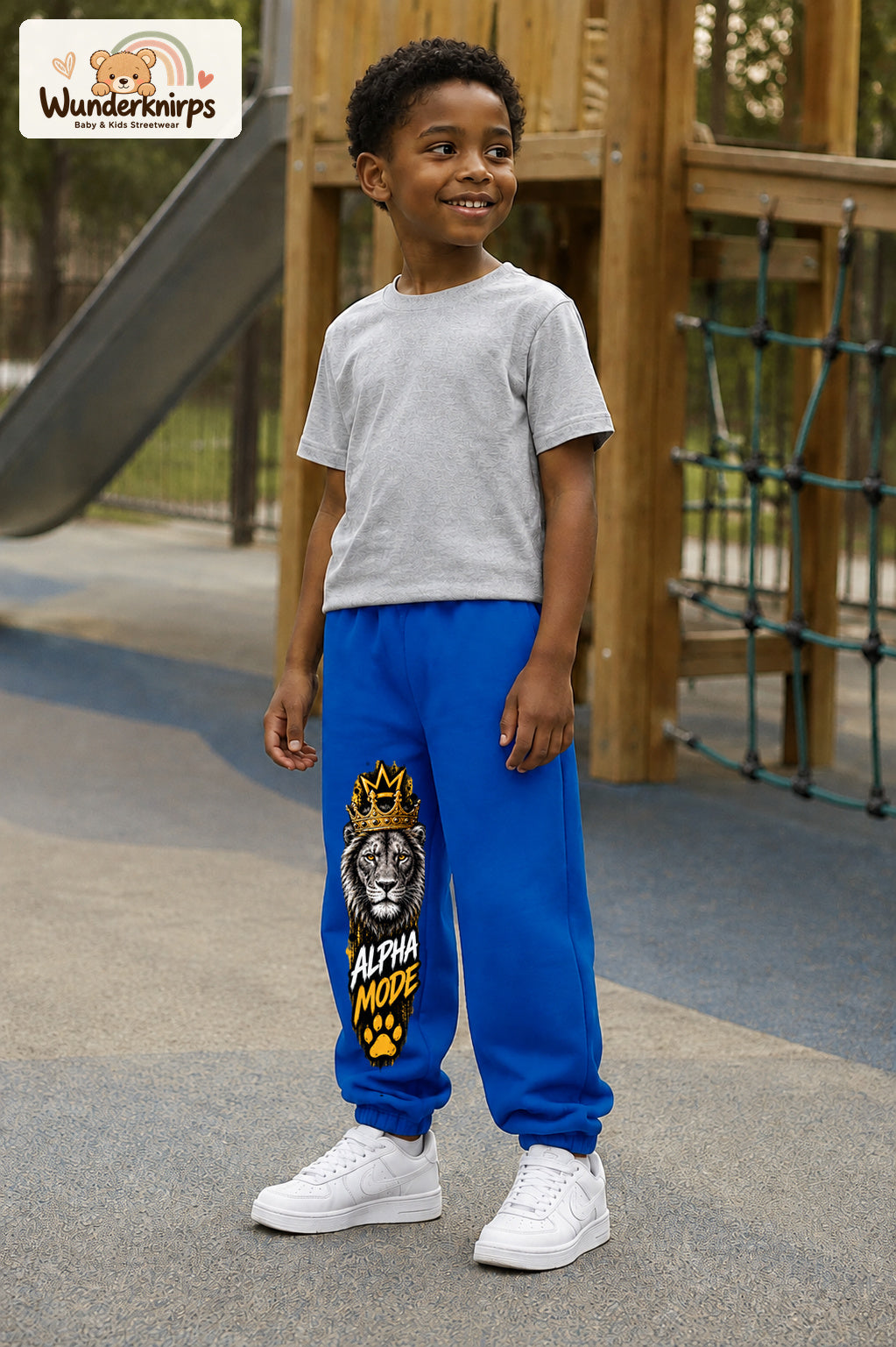 Junge trägt blaue Jogginghose mit Alpha Mode Löwe-Print, leicht seitlich auf dem Spielplatz.