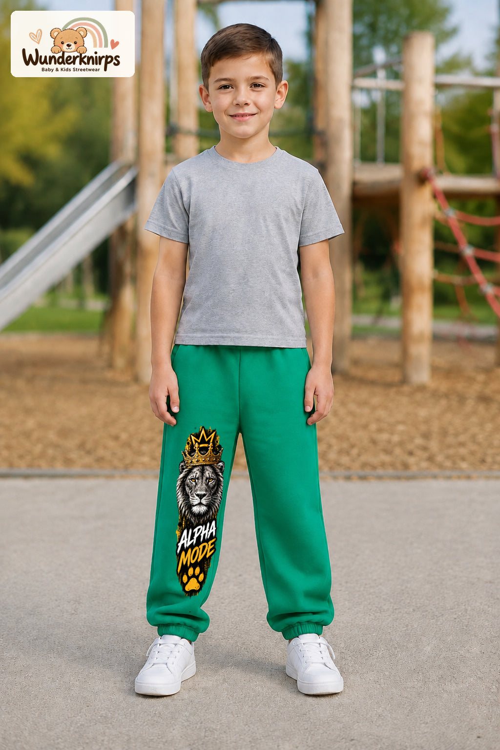 Junge trägt grüne Jogginghose mit Alpha Mode Löwe-Print, von vorn auf dem Spielplatz.