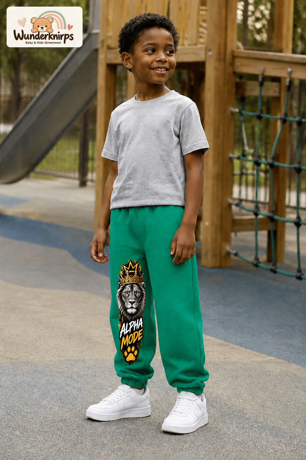 Junge trägt grüne Jogginghose mit Alpha Mode Löwe-Print, leicht seitlich auf dem Spielplatz.