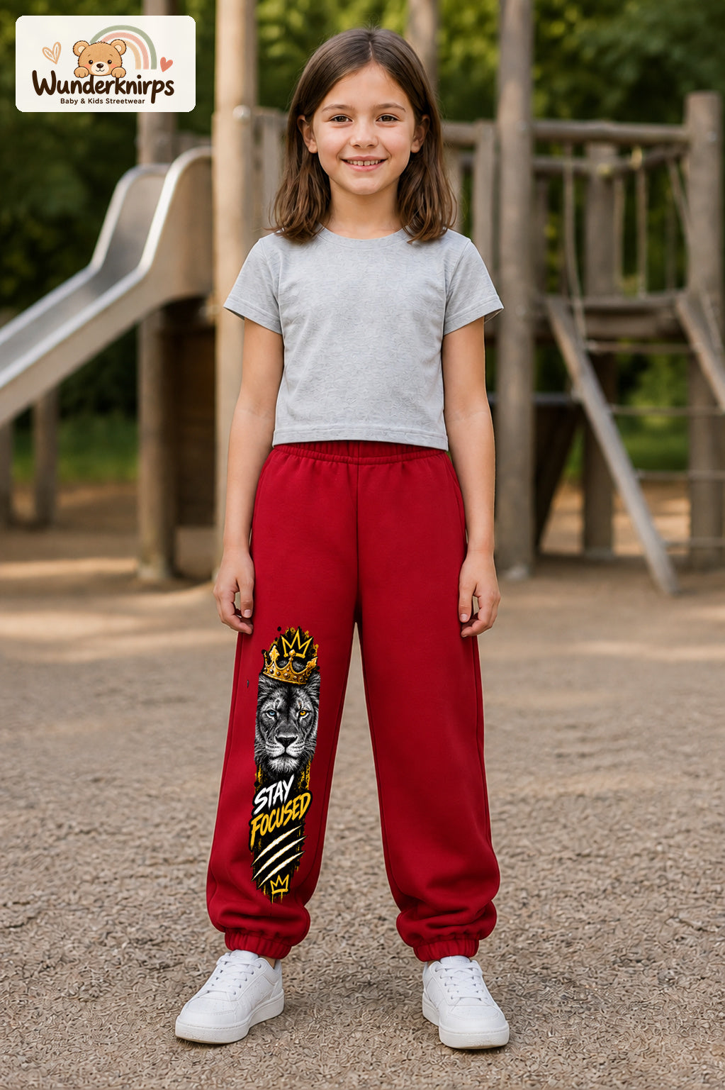 Mädchen trägt rote Jogginghose mit Stay Focused Löwe-Print, von vorn auf dem Spielplatz.