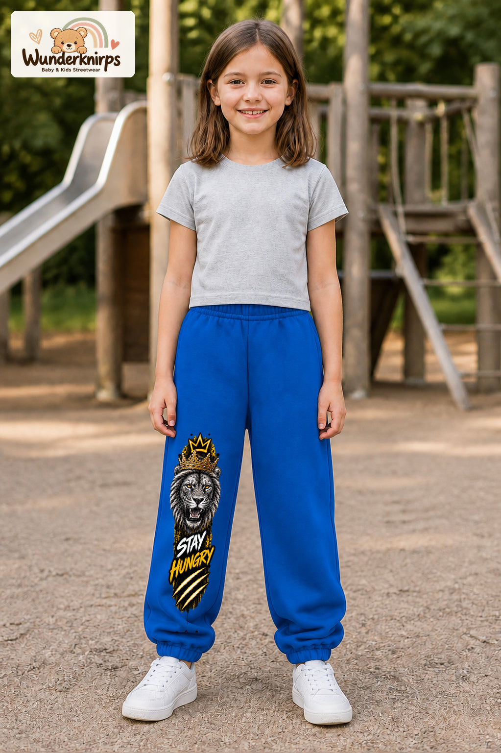 Mädchen trägt blaue Jogginghose mit Stay Hungry Löwe-Print, von vorn auf dem Spielplatz.