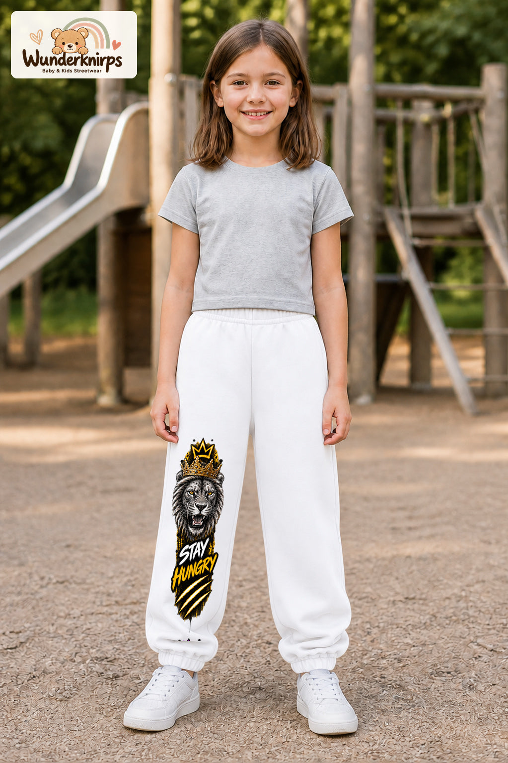 Mädchen trägt weiße Jogginghose mit Stay Hungry Löwe-Print, von vorn auf dem Spielplatz.