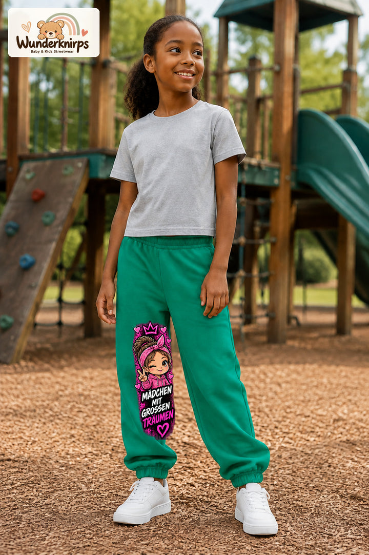 Mädchen trägt grüne Jogginghose mit Große Träume Herz-Bandana-Print, leicht seitlich auf dem Spielplatz.