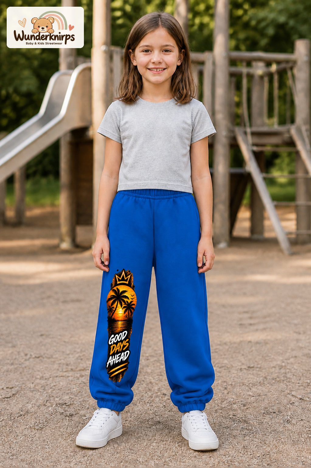 Mädchen trägt blaue Jogginghose mit Good Days Palmen-Print, von vorn auf dem Spielplatz.