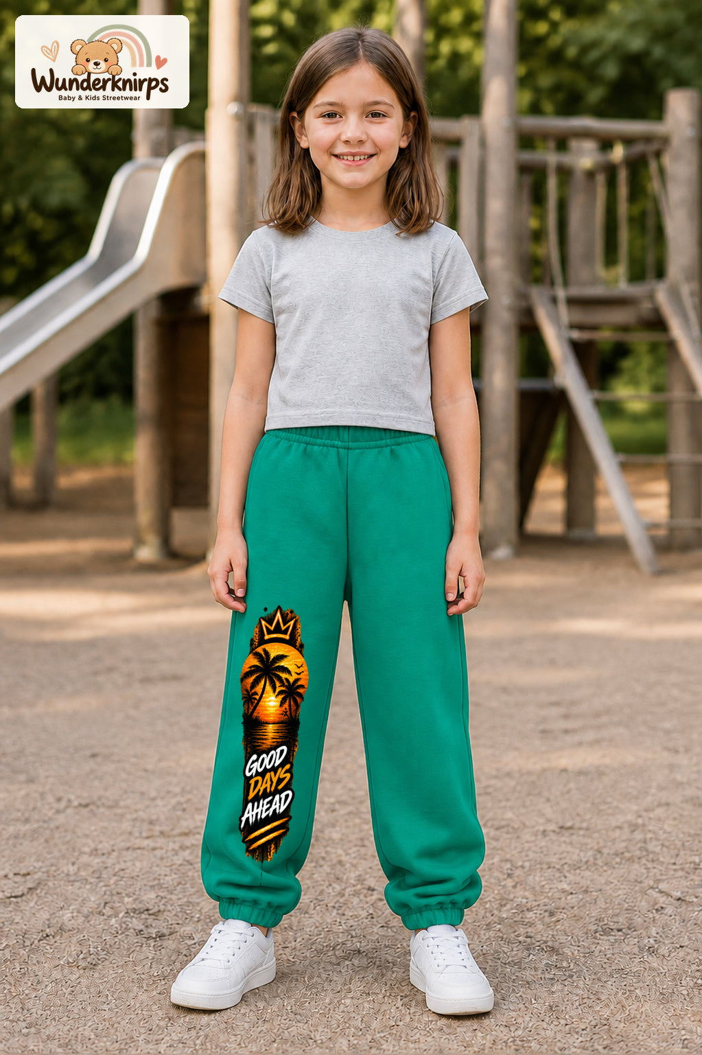 Mädchen trägt grüne Jogginghose mit Good Days Palmen-Print, von vorn auf dem Spielplatz.