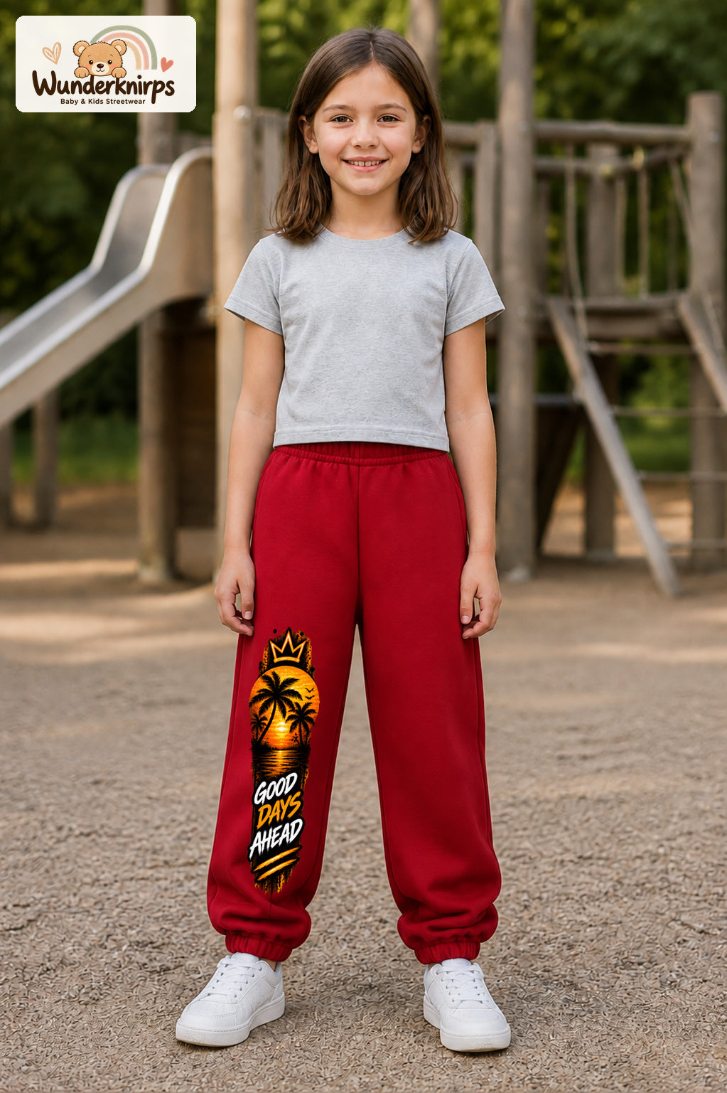 Mädchen trägt rote Jogginghose mit Good Days Palmen-Print, von vorn auf dem Spielplatz.