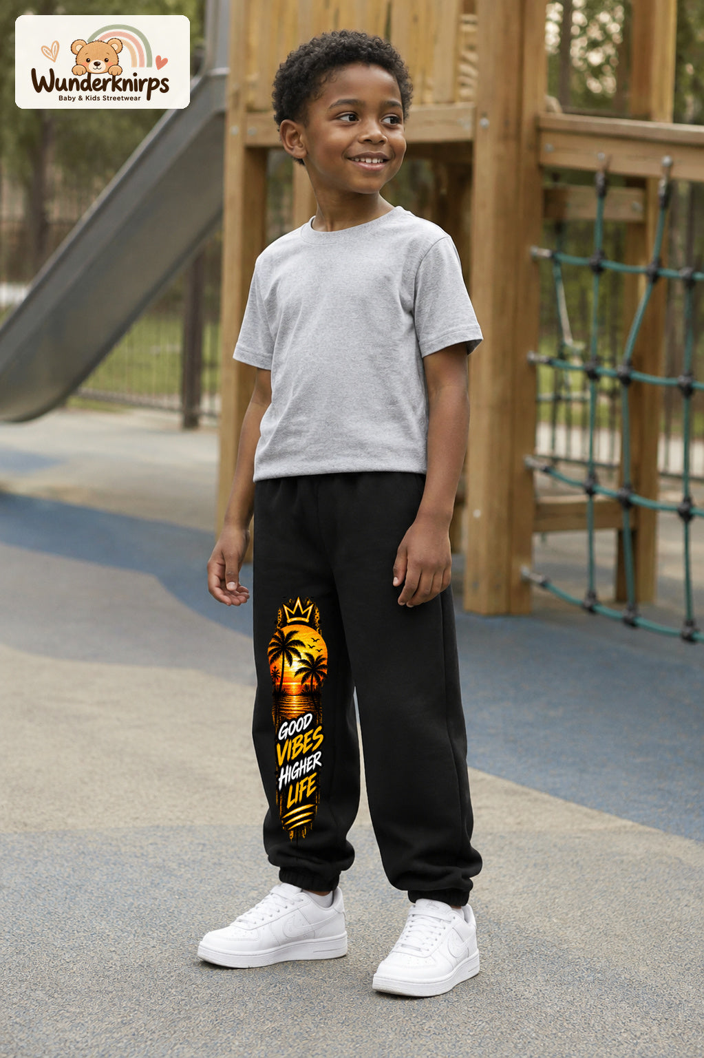 Junge trägt schwarze Jogginghose mit Higher Life Palmen-Print, leicht seitlich auf dem Spielplatz.