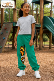 Mädchen trägt grüne Jogginghose mit Good Vibes Sonnenuntergang-Print, leicht seitlich auf dem Spielplatz.