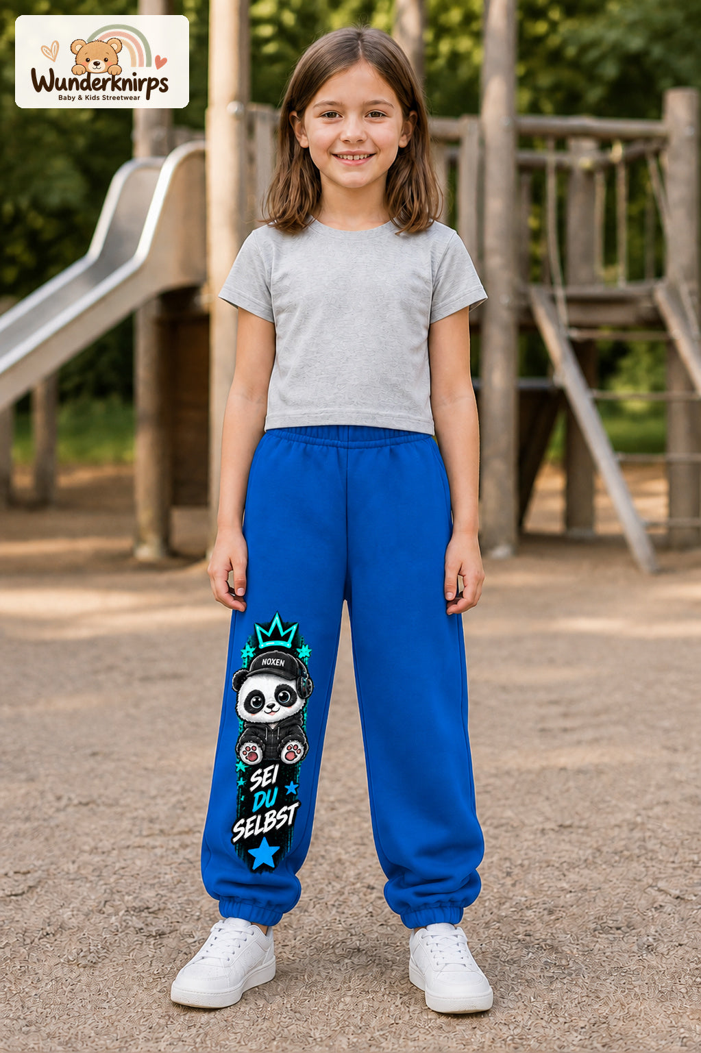 Mädchen trägt blaue Jogginghose mit Sei du selbst Panda-Print, von vorn auf dem Spielplatz.