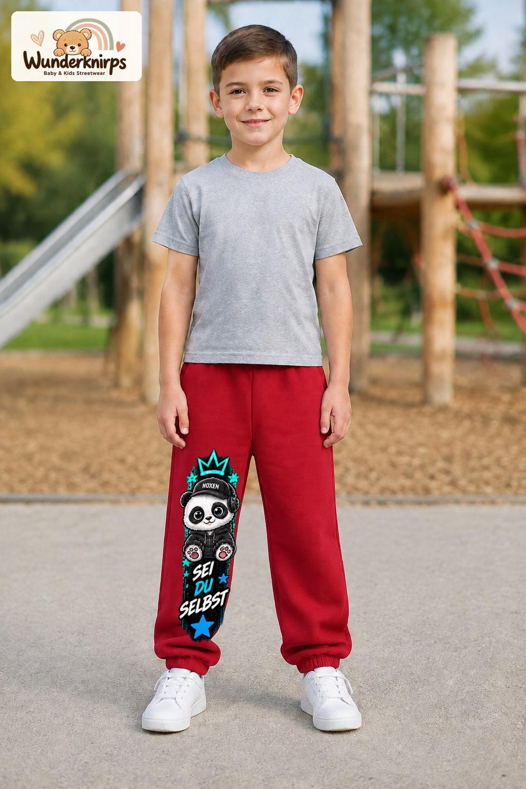 Junge trägt rote Jogginghose mit Sei du selbst Panda-Print, von vorn auf dem Spielplatz.