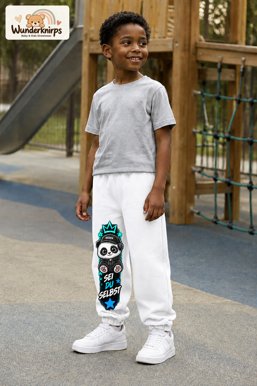 Junge trägt weiße Jogginghose mit Sei du selbst Panda-Print, leicht seitlich auf dem Spielplatz.
