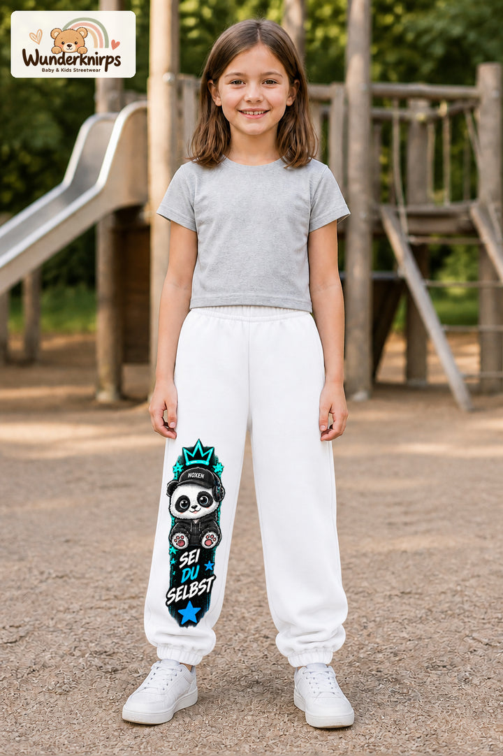 Mädchen trägt weiße Jogginghose mit Sei du selbst Panda-Print, von vorn auf dem Spielplatz.