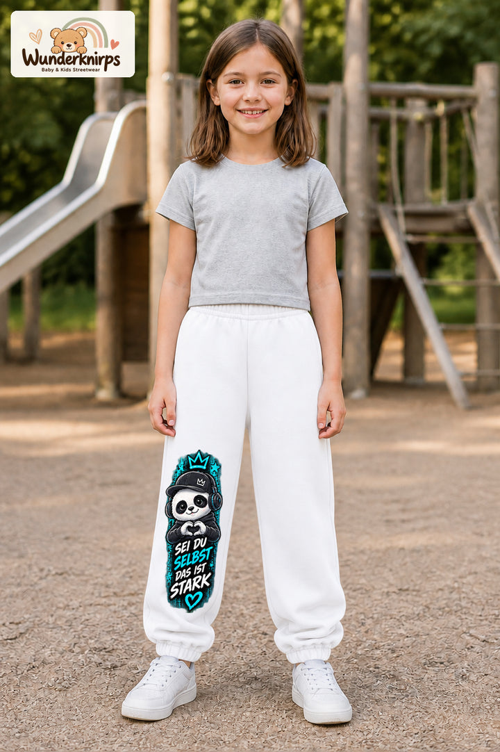 Mädchen trägt weiße Jogginghose mit Sei du selbst Herz-Panda-Print, von vorn auf dem Spielplatz.