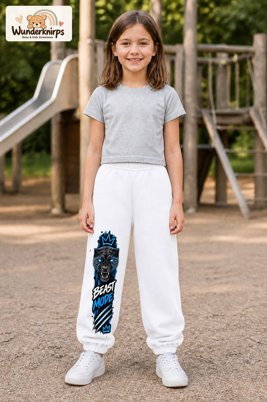 Mädchen trägt weiße Jogginghose mit Beast Mode Panther-Print, von vorn auf dem Spielplatz.