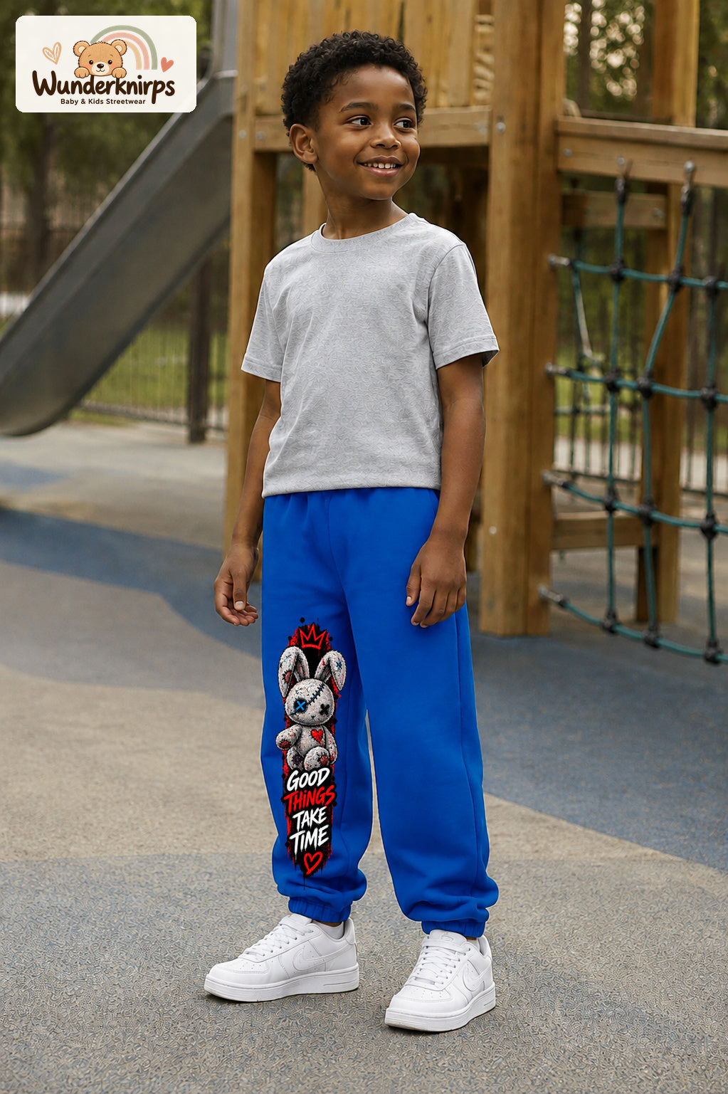 Junge trägt blaue Jogginghose mit Good Things Stoffhase-Print, leicht seitlich auf dem Spielplatz.