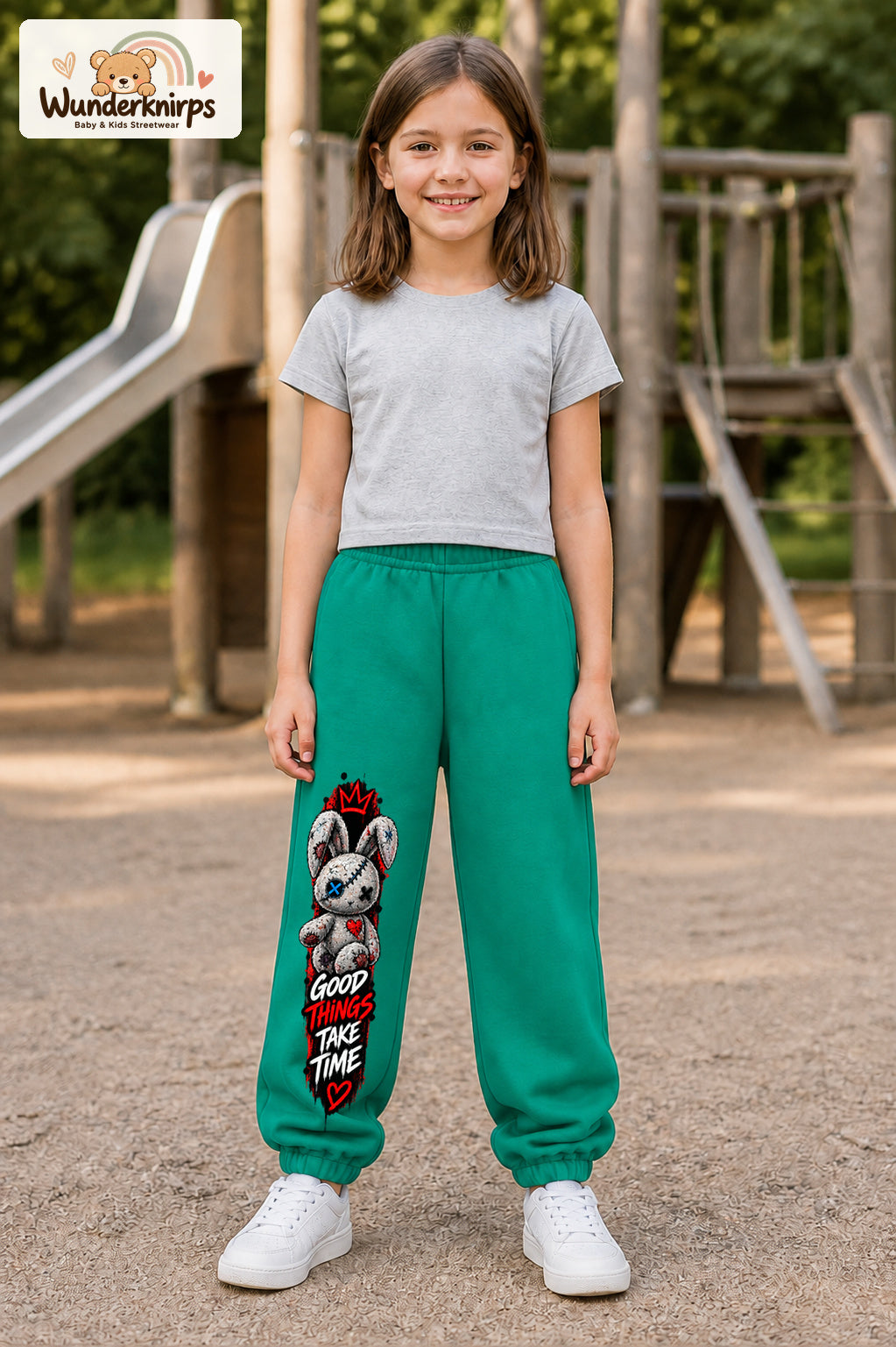 Mädchen trägt grüne Jogginghose mit Good Things Stoffhase-Print, von vorn auf dem Spielplatz.