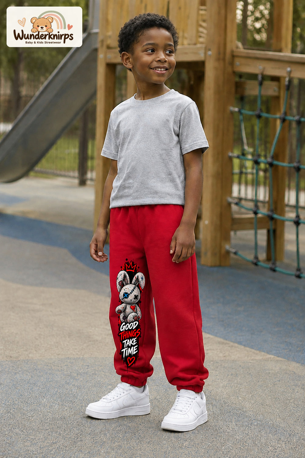 Junge trägt rote Jogginghose mit Good Things Stoffhase-Print, leicht seitlich auf dem Spielplatz.