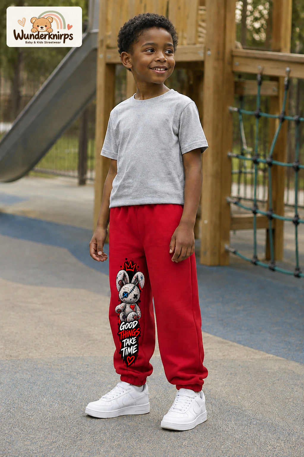 Junge trägt rote Jogginghose mit Good Things Stoffhase-Print, leicht seitlich auf dem Spielplatz.