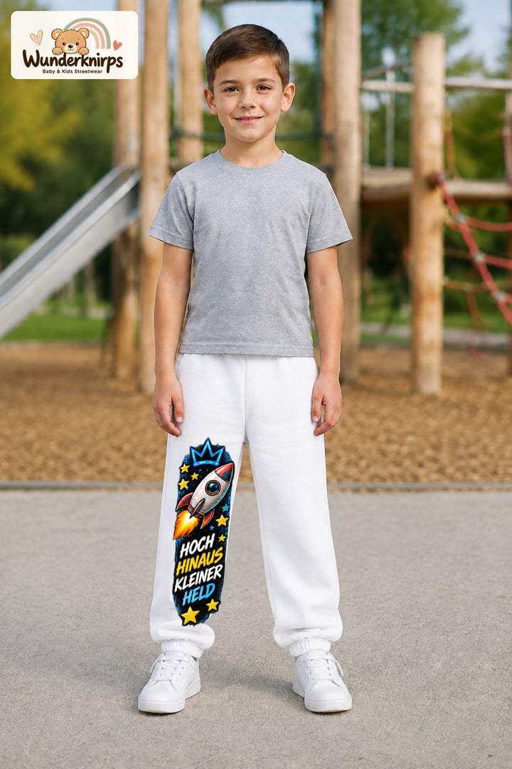 Junge trägt weiße Jogginghose mit Hoch hinaus Rakete-Print, von vorn auf dem Spielplatz.
