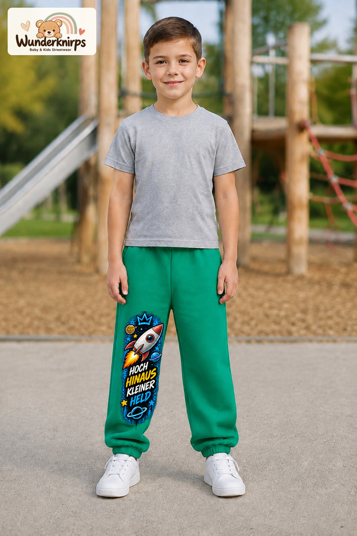 Junge trägt grüne Jogginghose mit Kleiner Held Weltraumrakete-Print, von vorn auf dem Spielplatz.